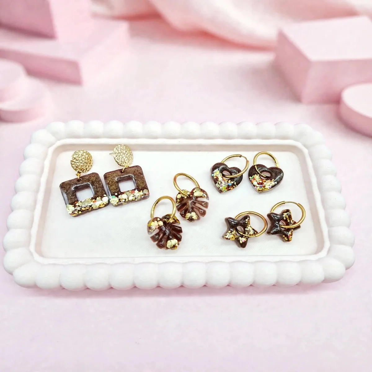 Boucles d'oreilles en résine marron et paillettes dorées : carrées, feuille de monstera, cœur et étoile, sur un plateau rose
