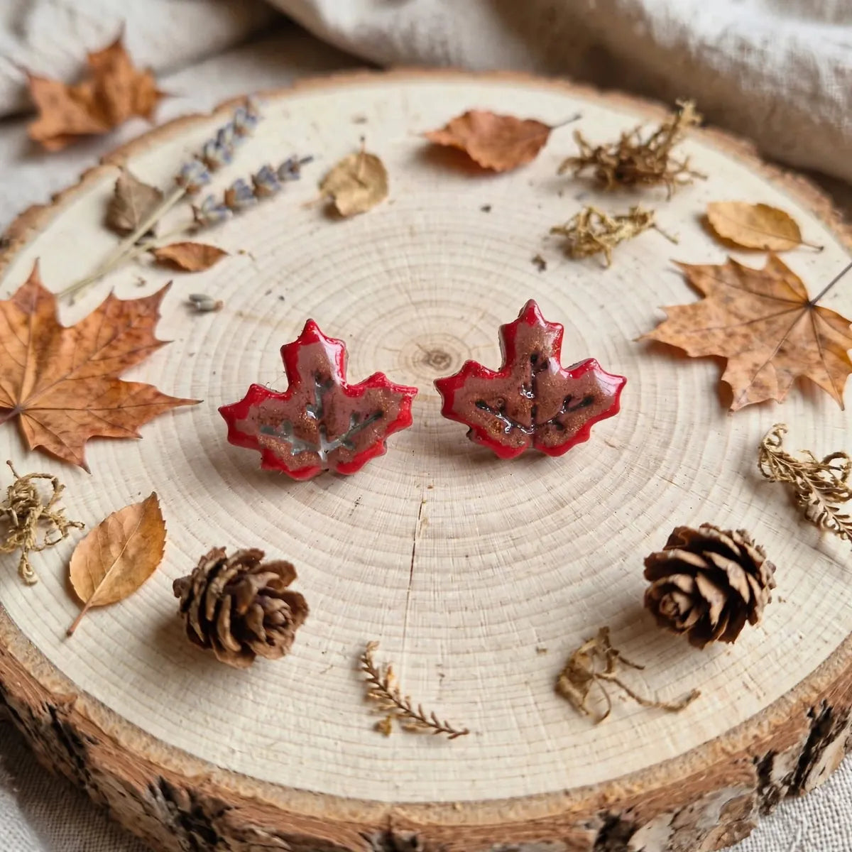 Paire de puces d’oreilles feuille d’érable rouge posée sur un rondin de bois parmi des feuilles et des pommes de pin
