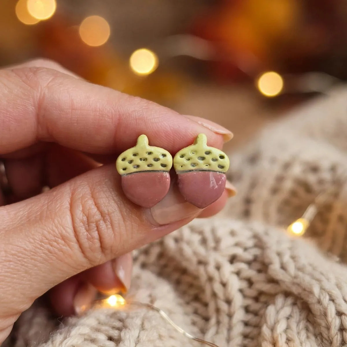 Deux puces d’oreilles gland vert clair et terracotta tenues à la main devant un pull en maille beige