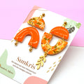 Boucles d'oreilles asymétriques fruit orange doré Sunkris sur carte de présentation, fond rose