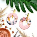 Boucles d'oreilles asymétriques arc rose doré Sunkris posées sur fond texturé, décor de fleurs séchées et coupelle terracotta