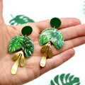 Main tenant la paire de boucles d'oreilles demi-lune monstera vert doré Sunkris, fond végétal