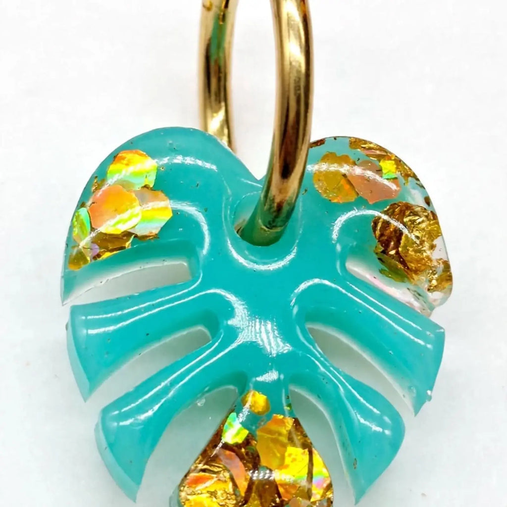Gros plan macro sur une boucle d'oreille feuille de monstera turquoise et paillettes dorées, anneau doré