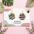 Boucles d'oreilles fleur rayée vert et rose Sunkris présentées sur socle rose entouré de fleurs séchées