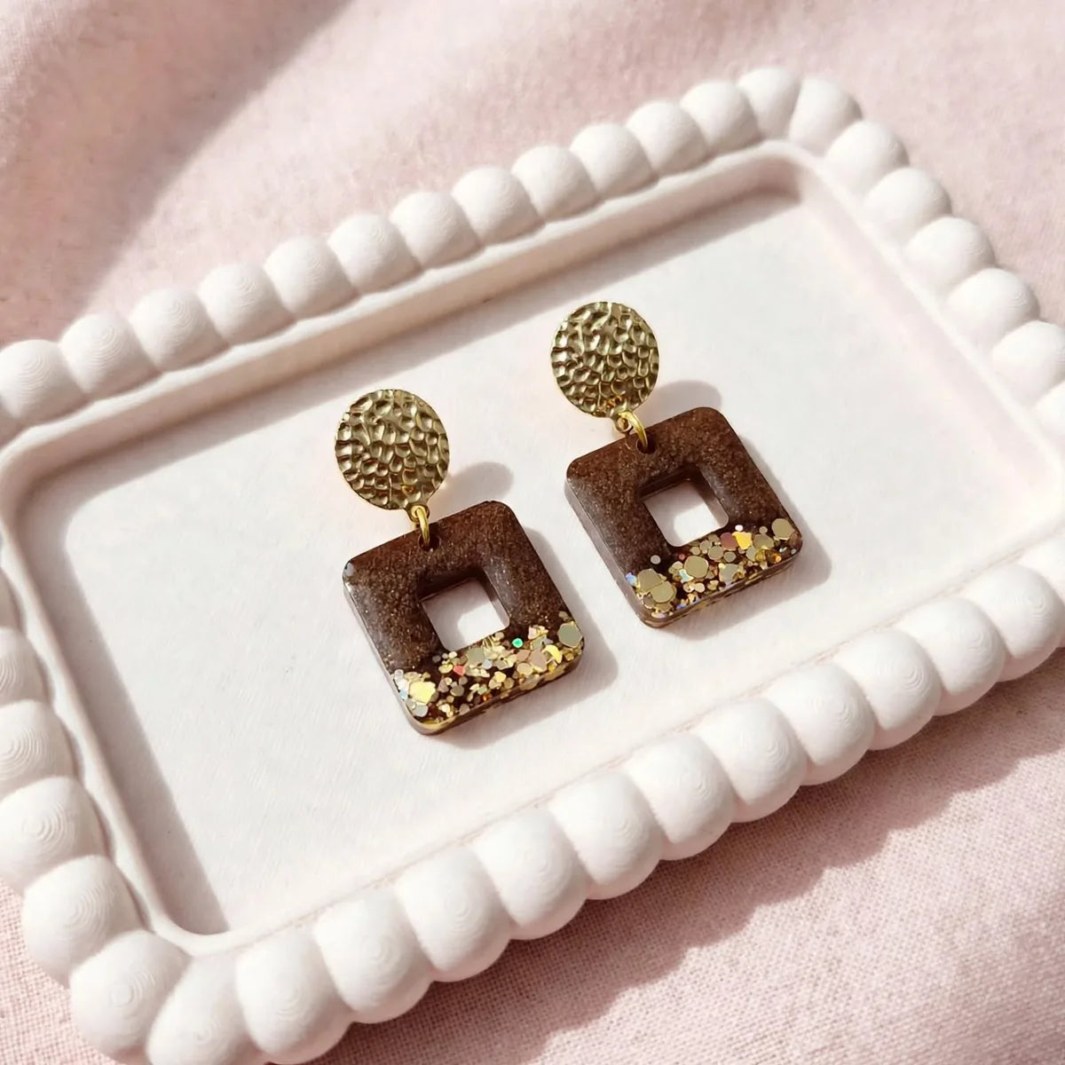 Boucles d'oreilles carrées en résine marron et paillettes dorées, puces dorées martelées, sur un plateau 
