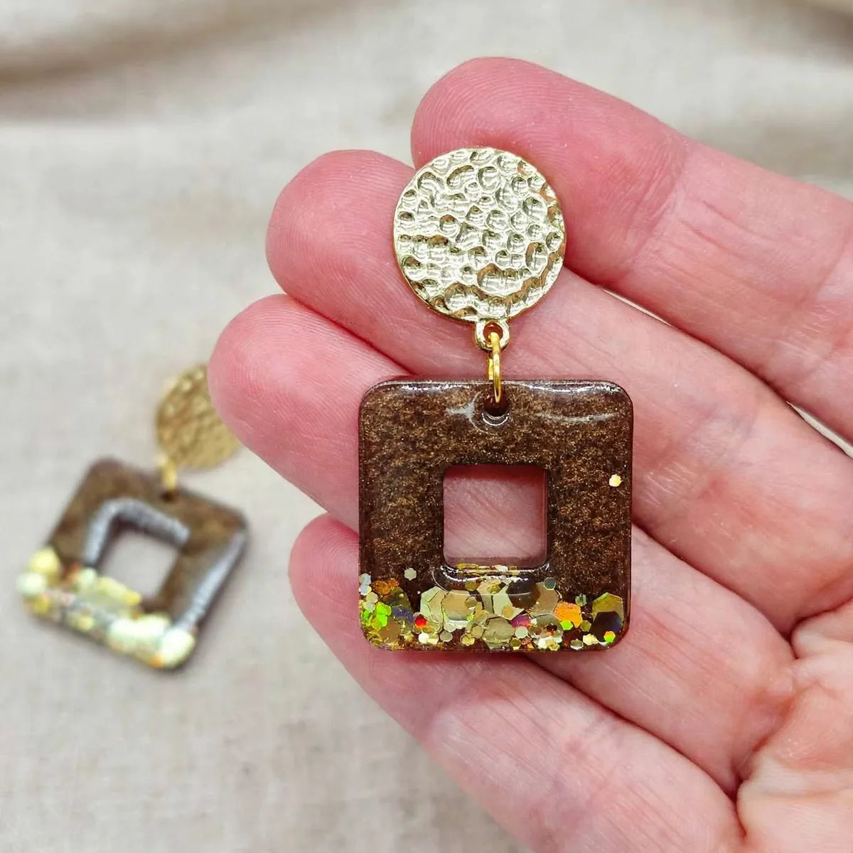 Boucle d'oreille carrée en résine marron et paillettes dorées tenue dans la main, seconde boucle en arrière-plan