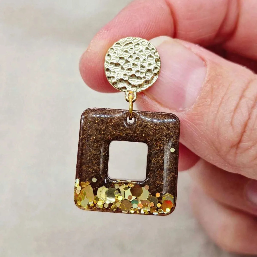 Gros plan d'une boucle d'oreille carrée en résine marron et paillettes dorées holographiques, puce dorée martelée