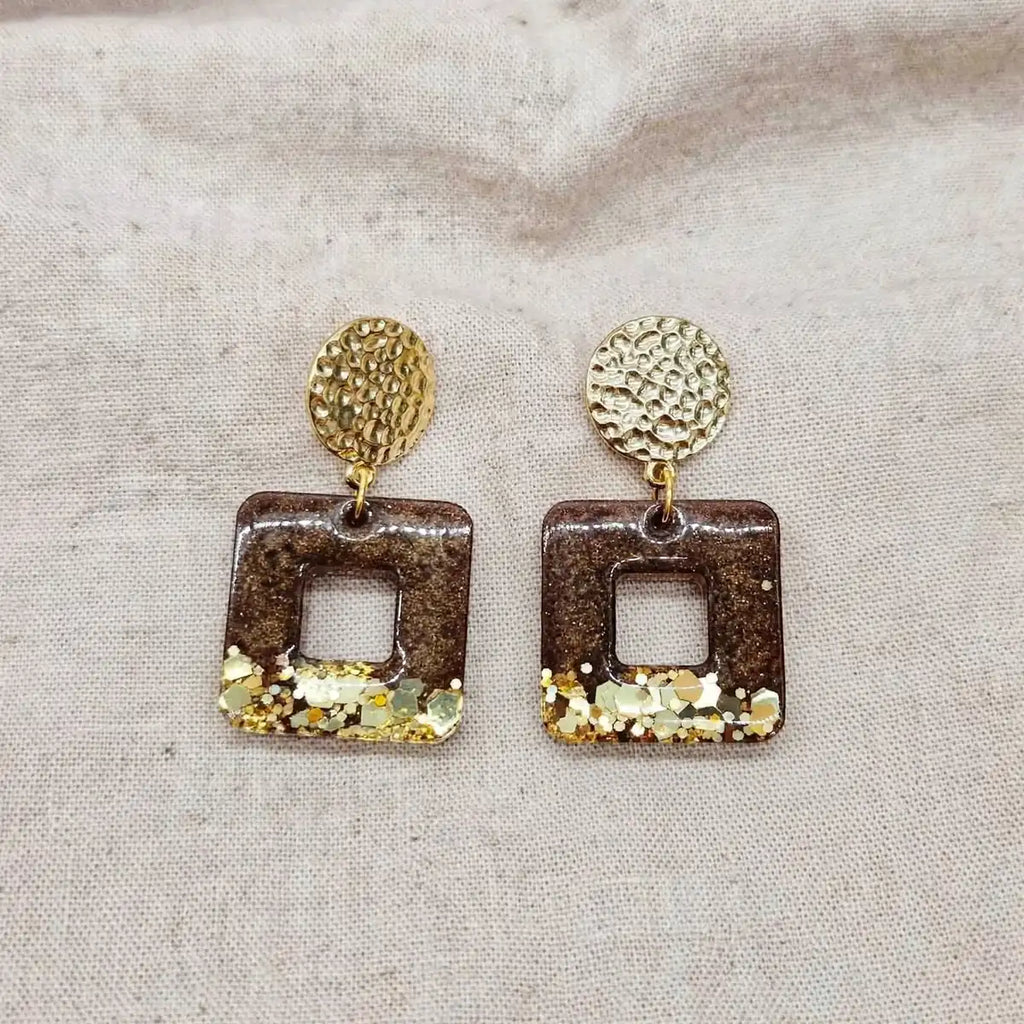 Paire de boucles d'oreilles carrées en résine marron et paillettes dorées sur fond de lin beige