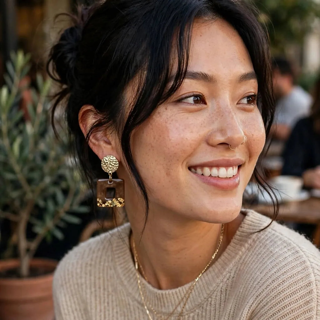 Femme souriante portant des boucles d'oreilles carrées en résine marron et paillettes dorées, en terrasse de café