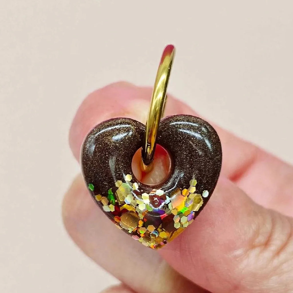 Gros plan d'une boucle d'oreille cœur en résine marron et paillettes holographiques tenue entre les doigts