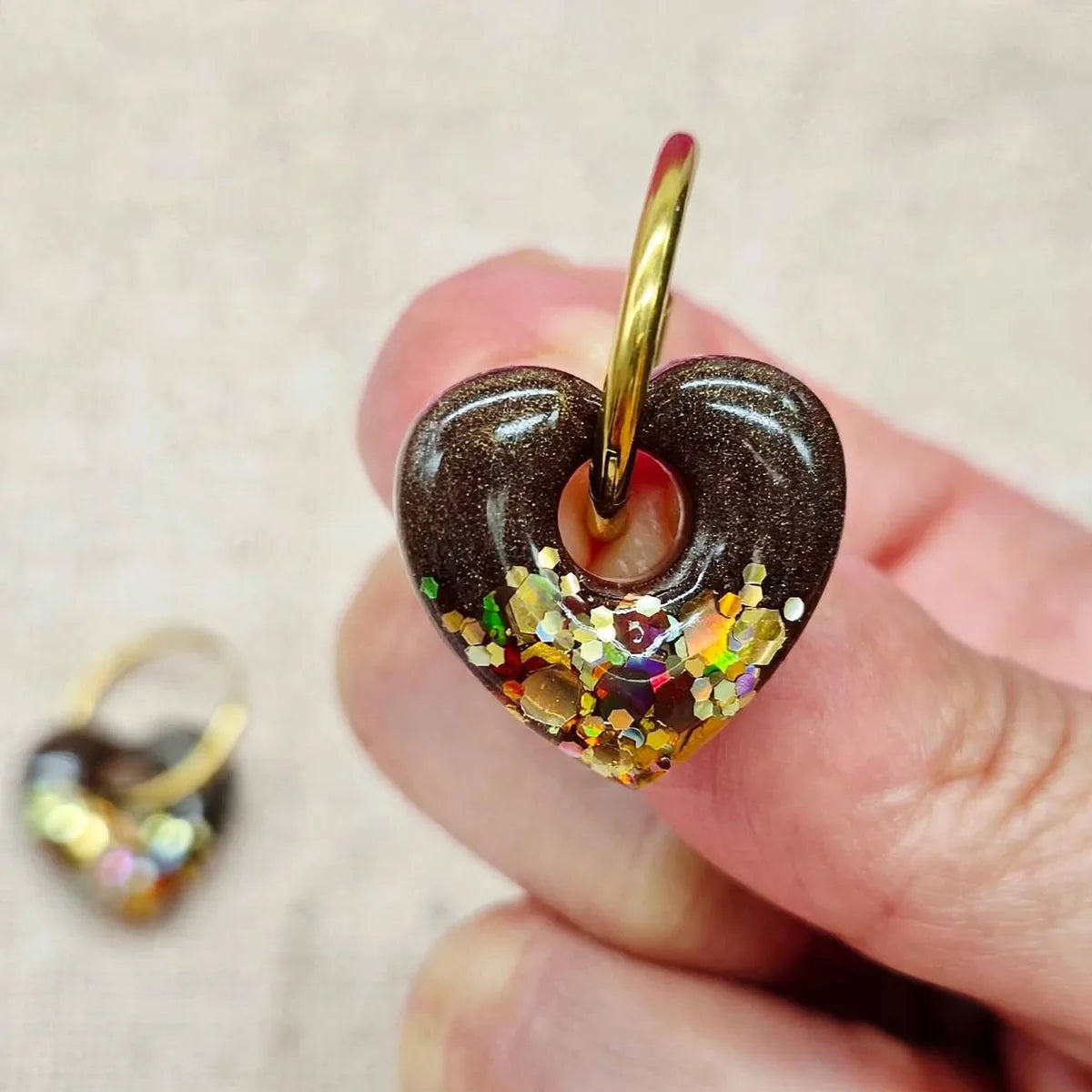 Boucle d'oreille cœur en résine marron et paillettes dorées tenue dans la main, seconde boucle floue au fond