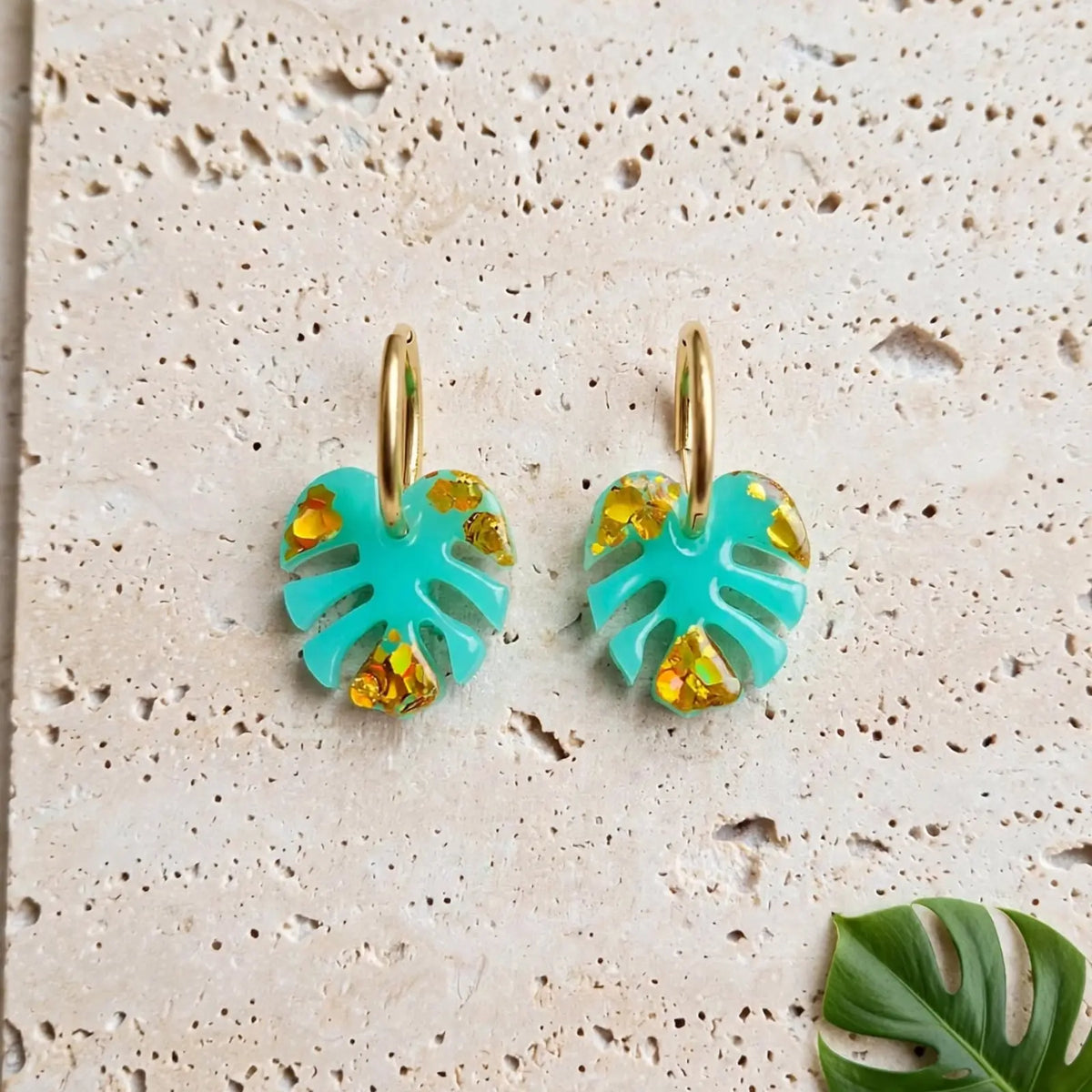 Boucles d'oreilles créoles dorées feuille de monstera turquoise posées sur pierre de travertin