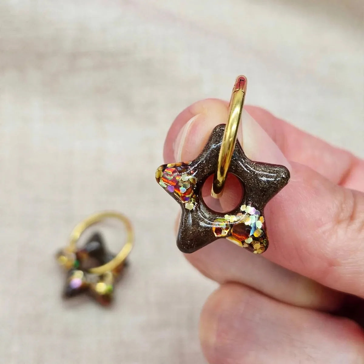 Boucle d'oreille étoile en résine marron et paillettes dorées tenue dans la main, seconde boucle floue au fond
