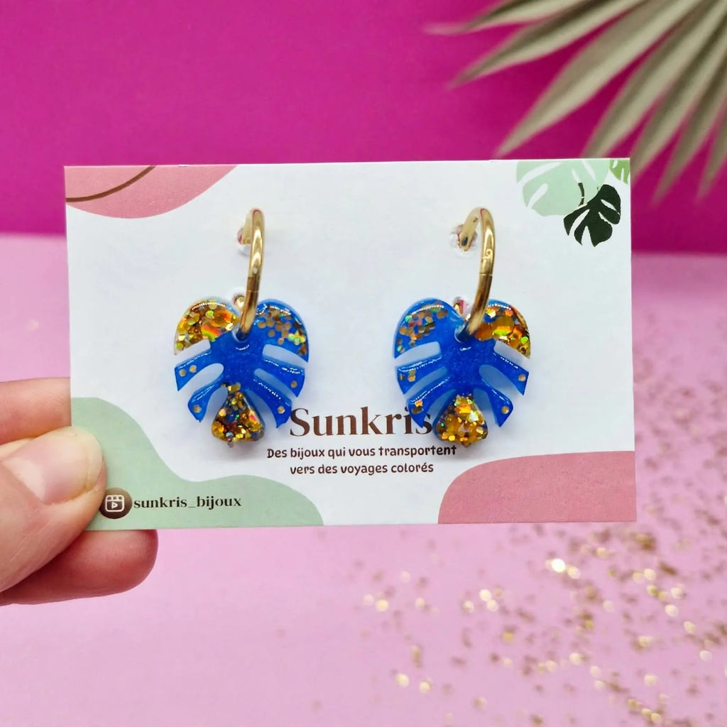 Boucles d'oreilles feuille de monstera bleu et or Sunkris présentées sur leur carte de marque