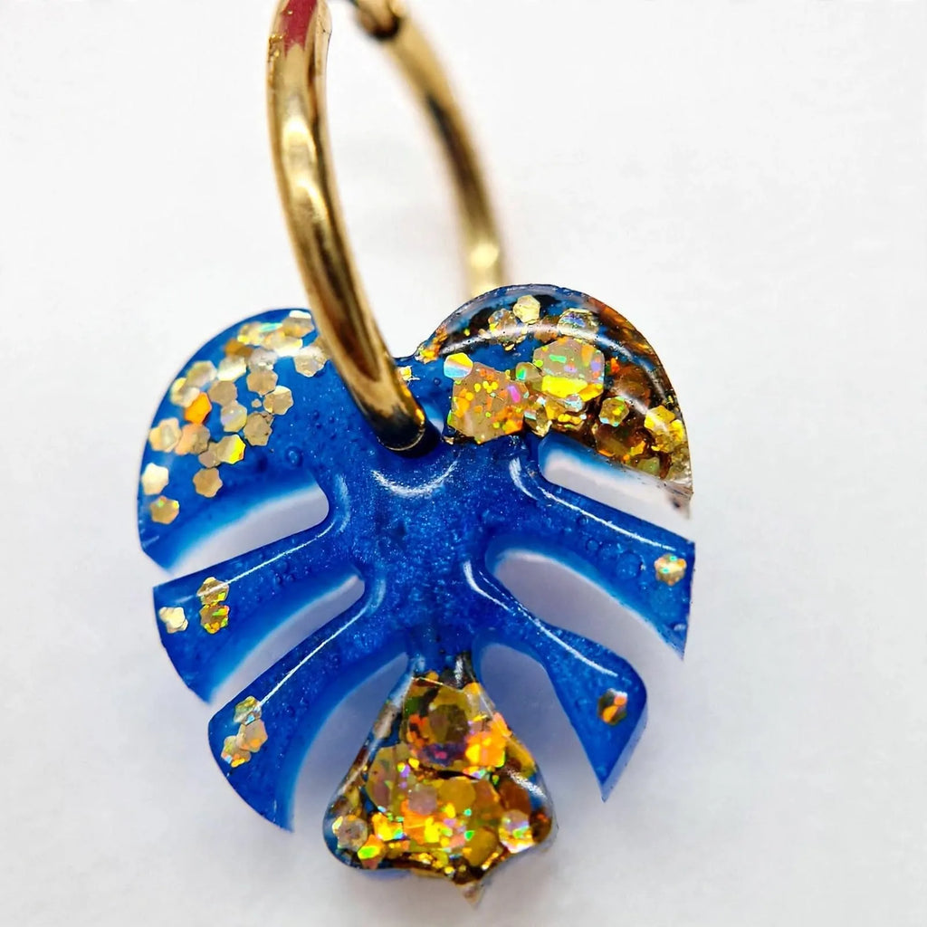 Gros plan sur une boucle d'oreille feuille de monstera en résine bleue pailletée et paillettes dorées Sunkris