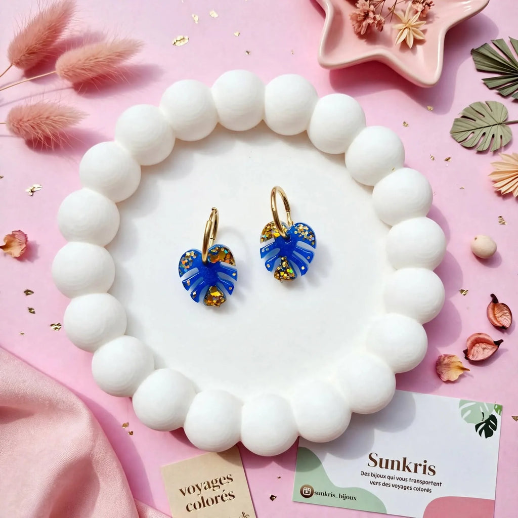 Boucles d'oreilles feuille de monstera bleu et or Sunkris posées sur un bracelet de perles blanches, mise en scène rose