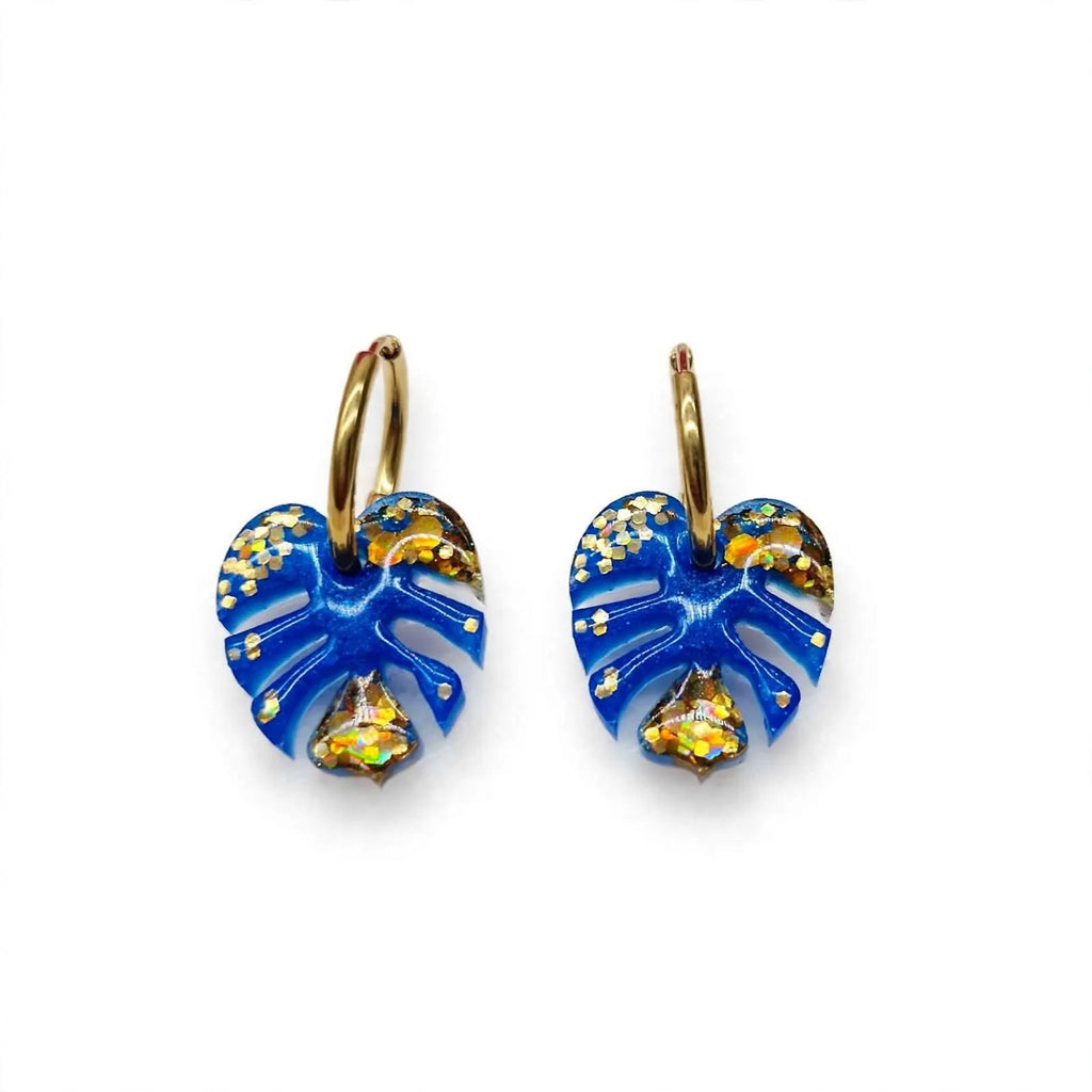 Boucles d'oreilles créoles dorées feuille de monstera en résine bleue pailletée sur fond blanc