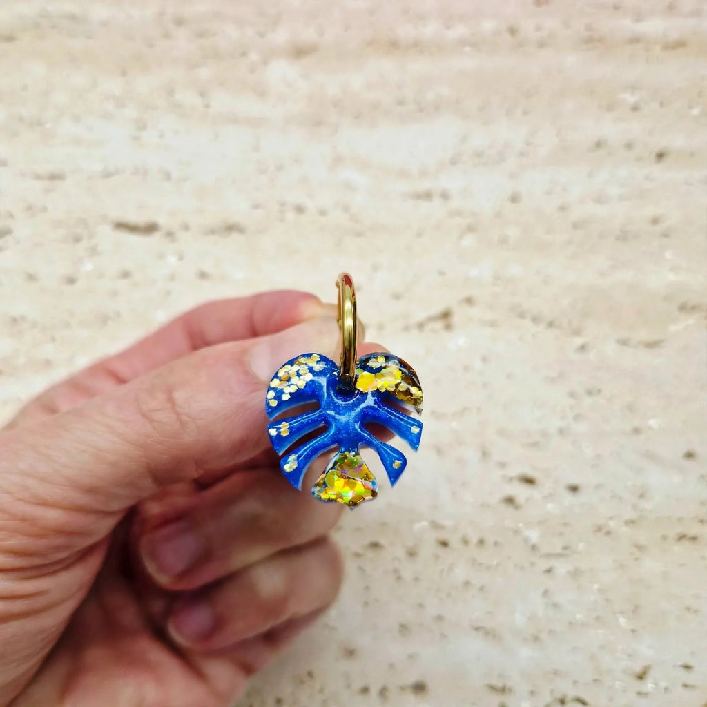 Boucle d'oreille feuille de monstera bleu et or Sunkris tenue en main, fond sable