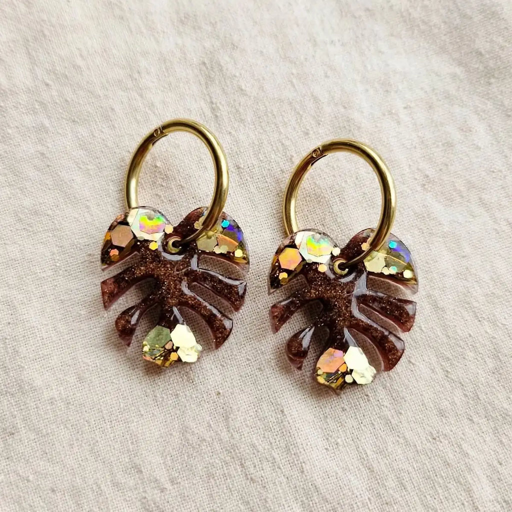 Paire de boucles d'oreilles feuille de monstera en résine marron et paillettes dorées sur créoles dorées, fond de lin