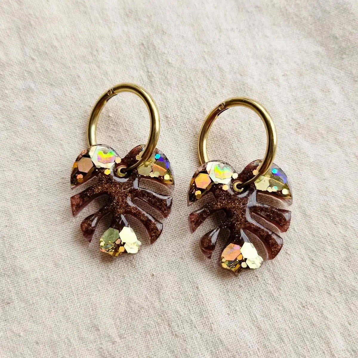 Paire de boucles d'oreilles feuille de monstera en résine marron et paillettes dorées sur créoles dorées, fond de lin