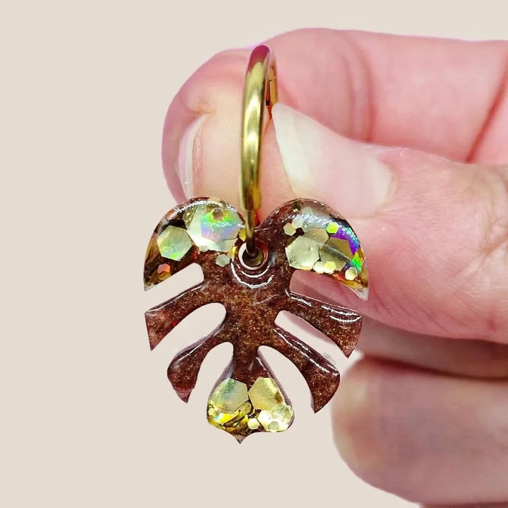 Gros plan d'une boucle d'oreille feuille de monstera en résine marron et paillettes dorées tenue entre les doigts