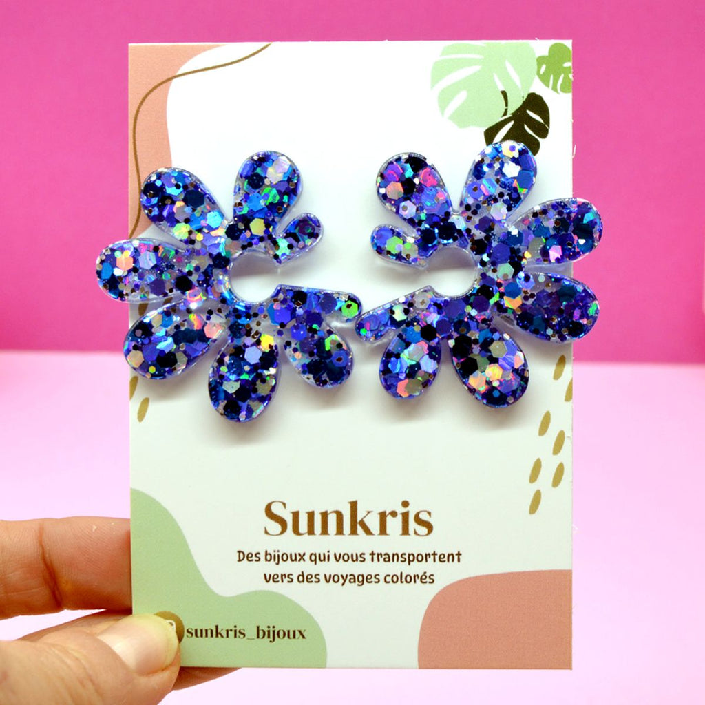 Boucles d’oreilles fleur à paillettes bleues sur leur carte Sunkris tenue en main, bijou artisanal en résine