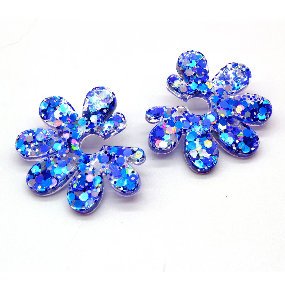 Boucles d’oreilles fleur en résine à paillettes bleues holographiques sur fond blanc, bijou artisanal Sunkris