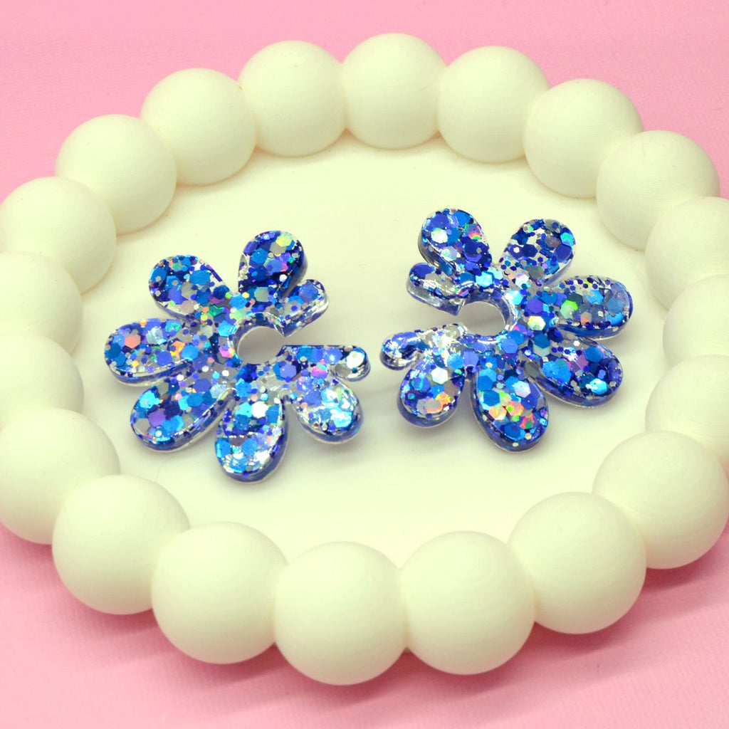 Boucles d’oreilles fleur paillettes bleu holographique en résine posées sur un plateau de perles blanches