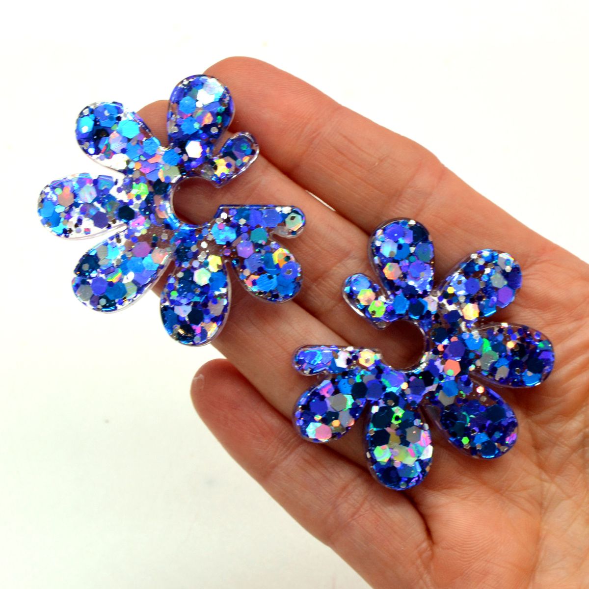 Boucles d’oreilles fleur en résine à paillettes bleues holographiques tenues dans la paume d’une main